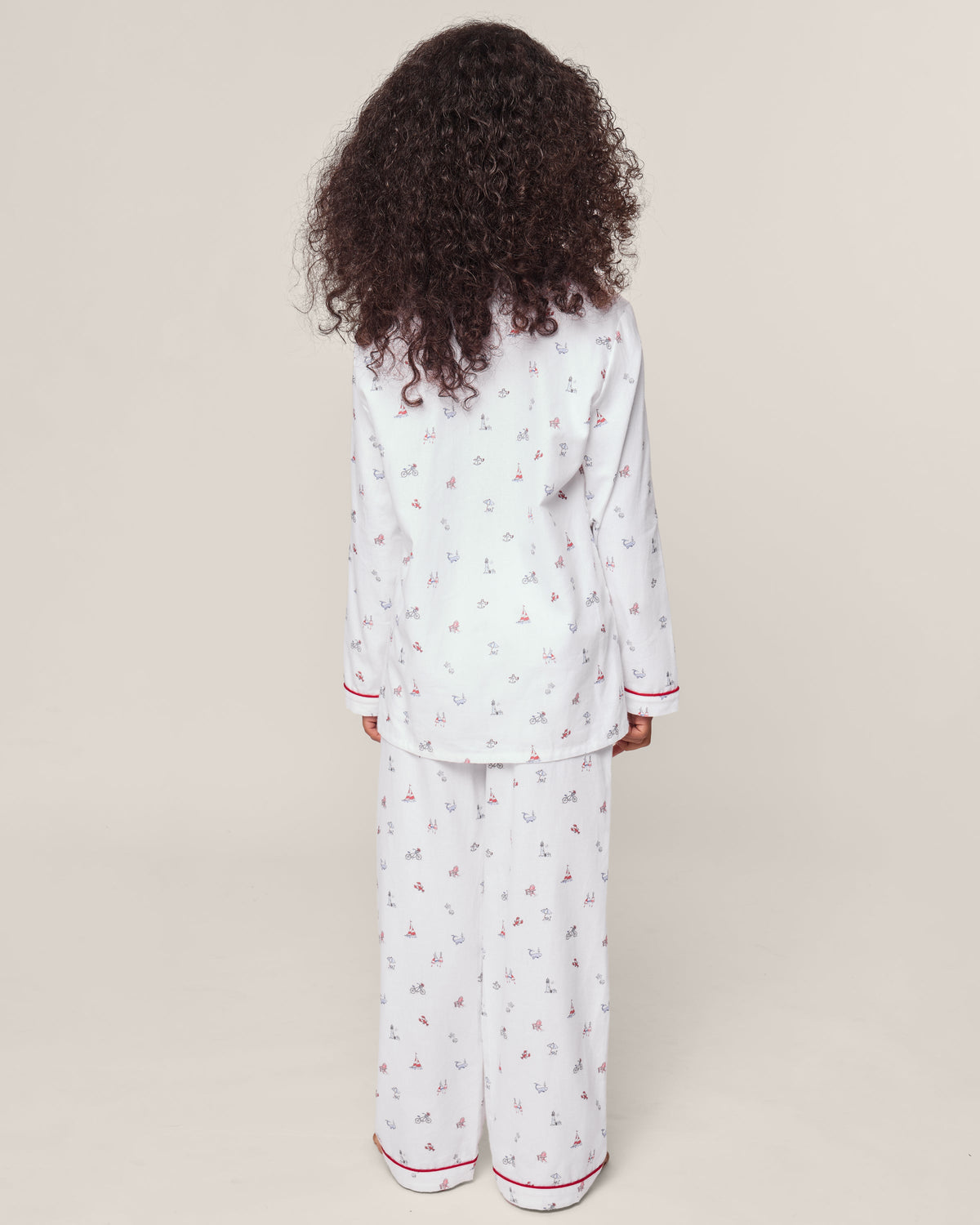 A child with curly hair, wearing a Petite Plume Kids Twill Pajama in Coastal Charms, featuring white yarn-dyed cotton and red and blue patterns, stands with their back to the camera against a plain background.