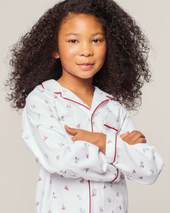 A curly-haired child grins with crossed arms, wearing Petite Plumes Kids Twill Pajama in Coastal Charms. The white cotton top showcases small red and blue patterns, accented by red piping, set against a light background.