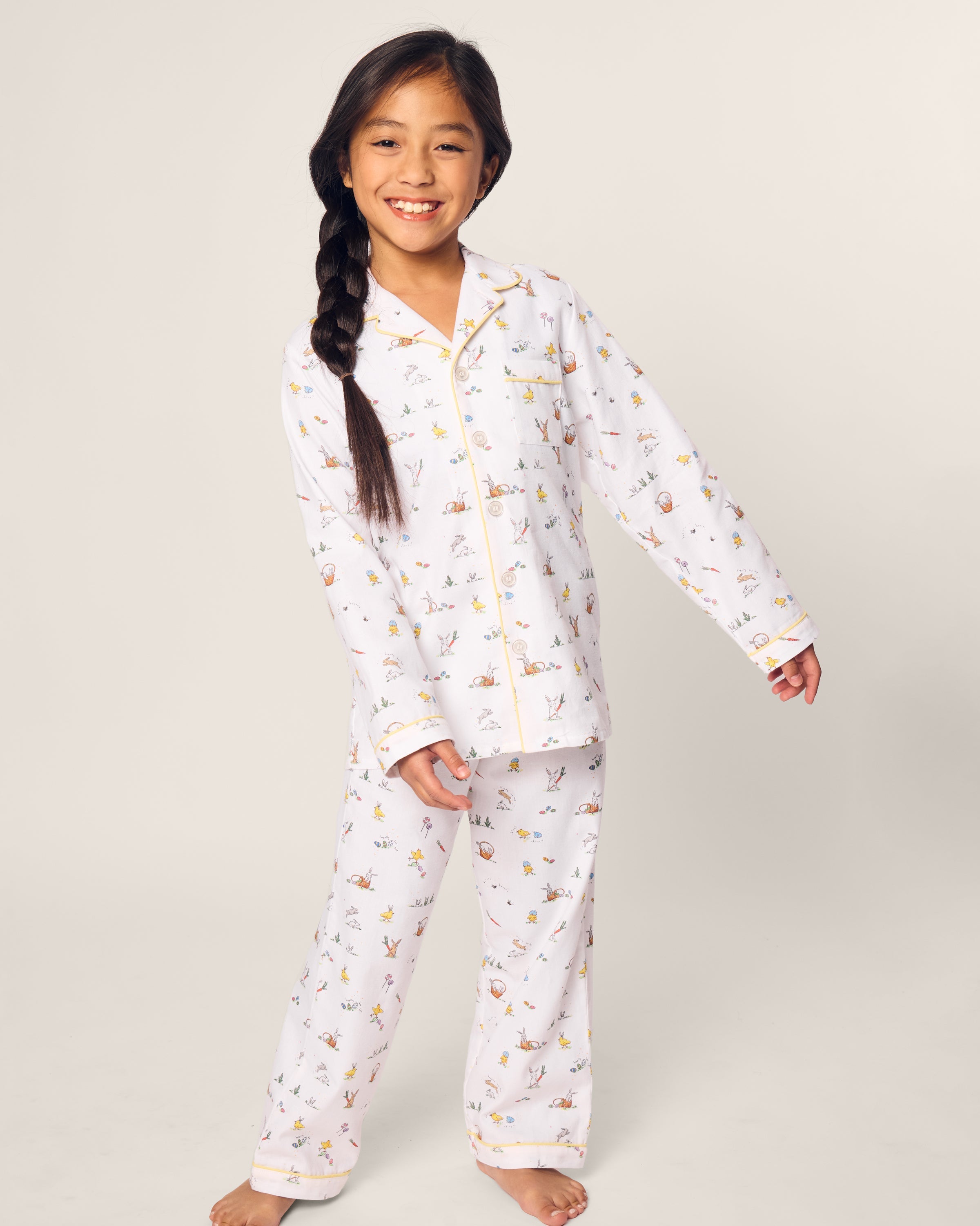 A young girl with long dark hair in a side braid rocks the Petite Plume Kids Twill Pajama in Easter Frolic, her smile beaming as she strikes a playful pose against a plain background.