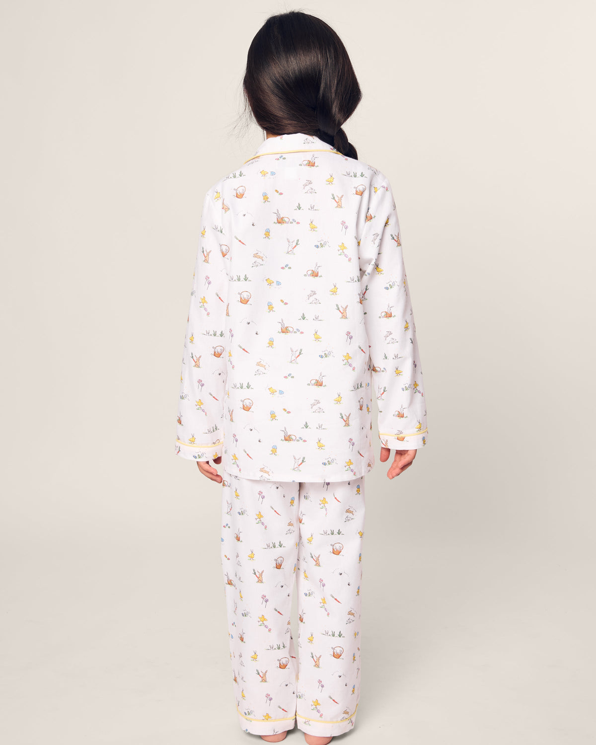 A child with long dark hair stands facing away in Petite Plumes Kids Twill Pajama, a white set adorned with pastel animal and balloon patterns ideal for Easter Frolic. This flame retardant sleepwear is charmingly displayed against a plain, light-colored background.