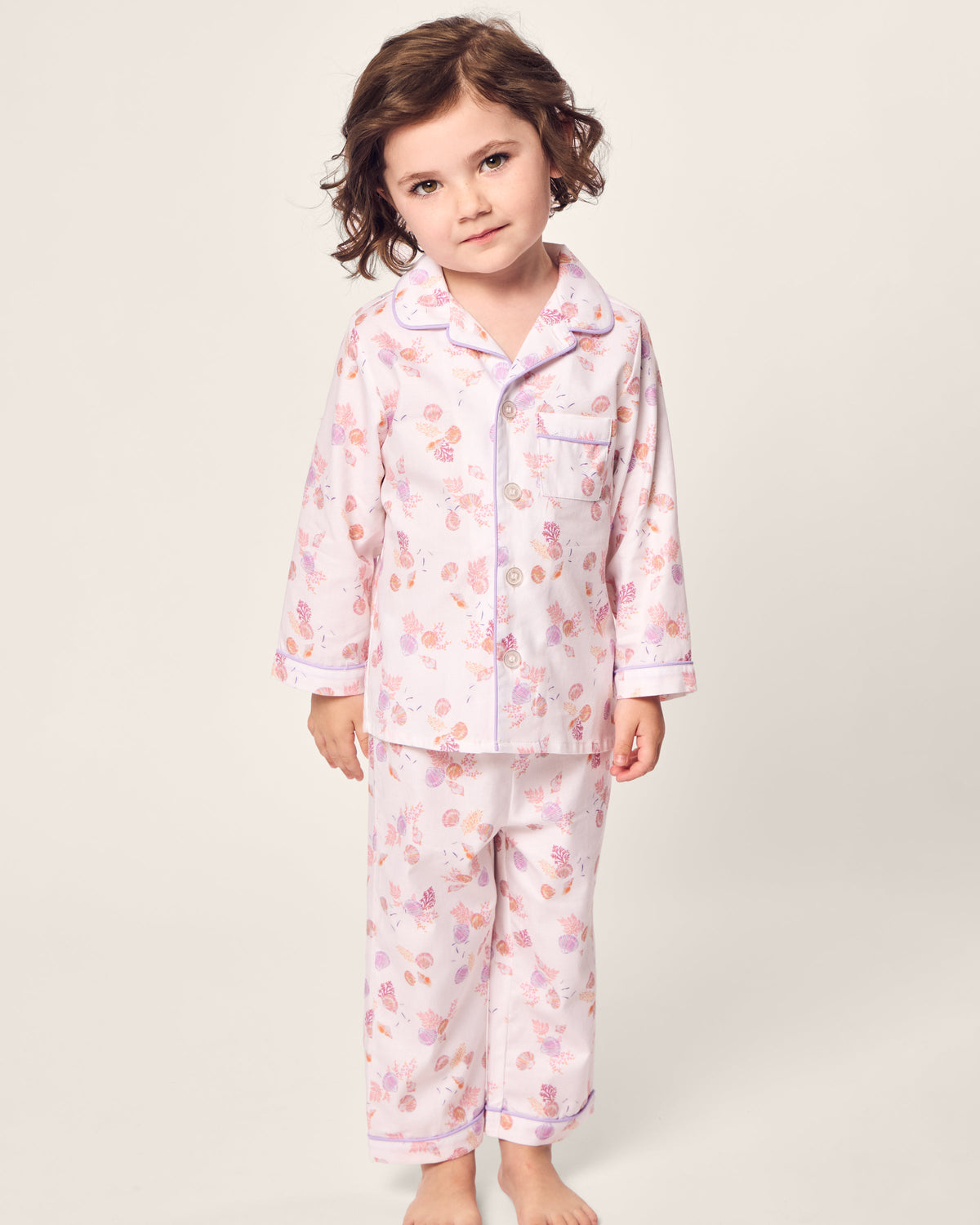 Amid a plain backdrop, a child with brown curly hair gazes to one side, wearing Petite Plumes Kids Twill Pajama in Seashell Splendor—a yarn-dyed cotton set with a pink and purple floral design. The collared button-up top and matching pants embody cozy charm.