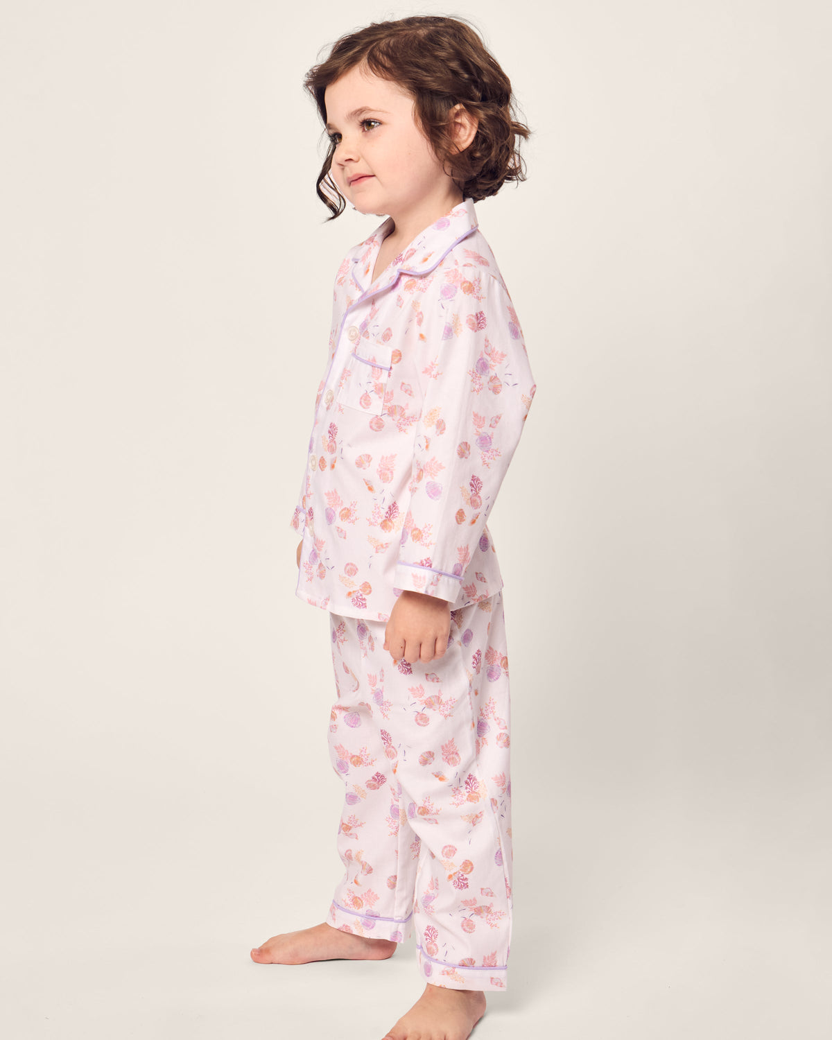 A young child with curly hair stands barefoot in Petite Plumes cozy Kids Twill Pajama in Seashell Splendor, featuring pink patterns on soft white cotton. This flame retardant sleepwear provides safety and comfort against a plain light background as they gaze into the distance, relaxed.
