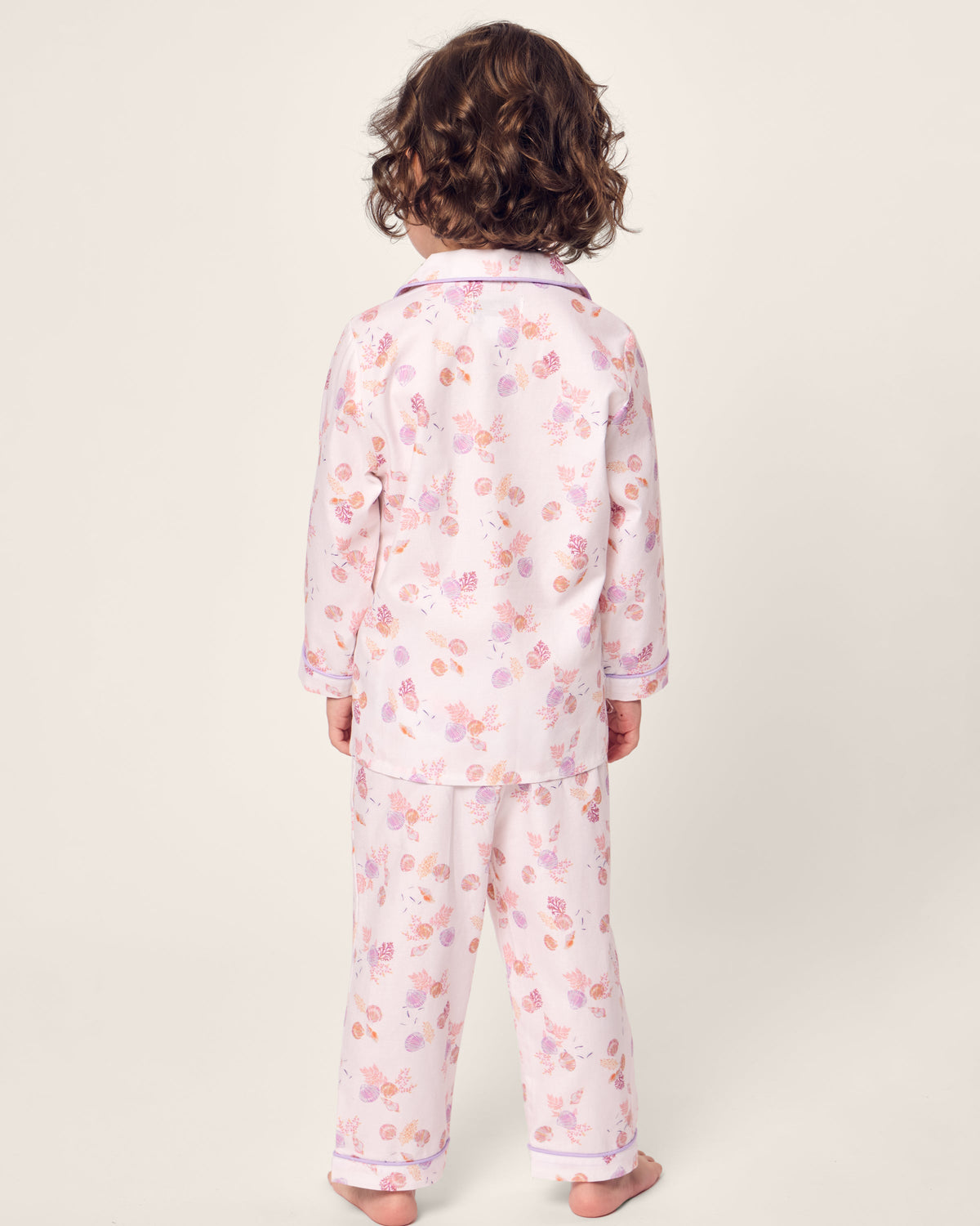 A young child with curly hair faces away from the camera, wearing the Petite Plume Kids Twill Pajama in Seashell Splendor, a flame retardant set made of yarn-dyed cotton. The pink pajamas display a charming floral pattern on a plain, light-hued background.