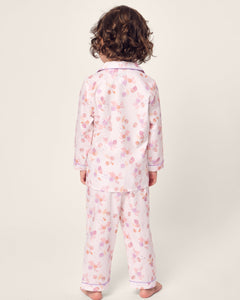 A young child with curly hair faces away from the camera, wearing the Petite Plume Kids Twill Pajama in Seashell Splendor, a flame retardant set made of yarn-dyed cotton. The pink pajamas display a charming floral pattern on a plain, light-hued background.