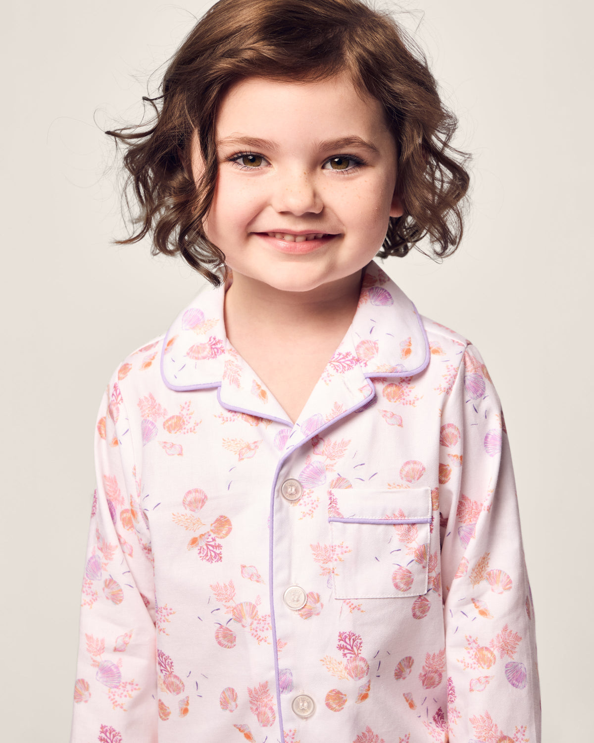 A young girl with short, curly brown hair smiles in Petite Plumes Kids Twill Pajama in Seashell Splendor. The charming yarn-dyed cotton pajamas with a pink and orange floral pattern, collar, and buttons stand out against a light, neutral background.