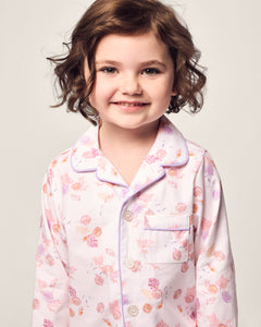 A young girl with short, curly brown hair smiles in Petite Plumes Kids Twill Pajama in Seashell Splendor. The charming yarn-dyed cotton pajamas with a pink and orange floral pattern, collar, and buttons stand out against a light, neutral background.