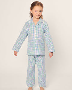 A young child poses barefoot wearing the Petite Plume Kids Twill Pajama in Spring Gingham, featuring light blue checks, yarn-dyed cotton fabric, a collar, and buttons, gently smiling at the camera against a plain background.