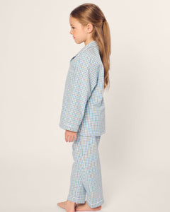 A young girl with long hair stands sideways, barefoot on a plain background, wearing the Petite Plume Kids Twill Pajama in Spring Gingham. Made from yarn-dyed cotton, it offers comfort and style with its cozy blue and white checkered design.