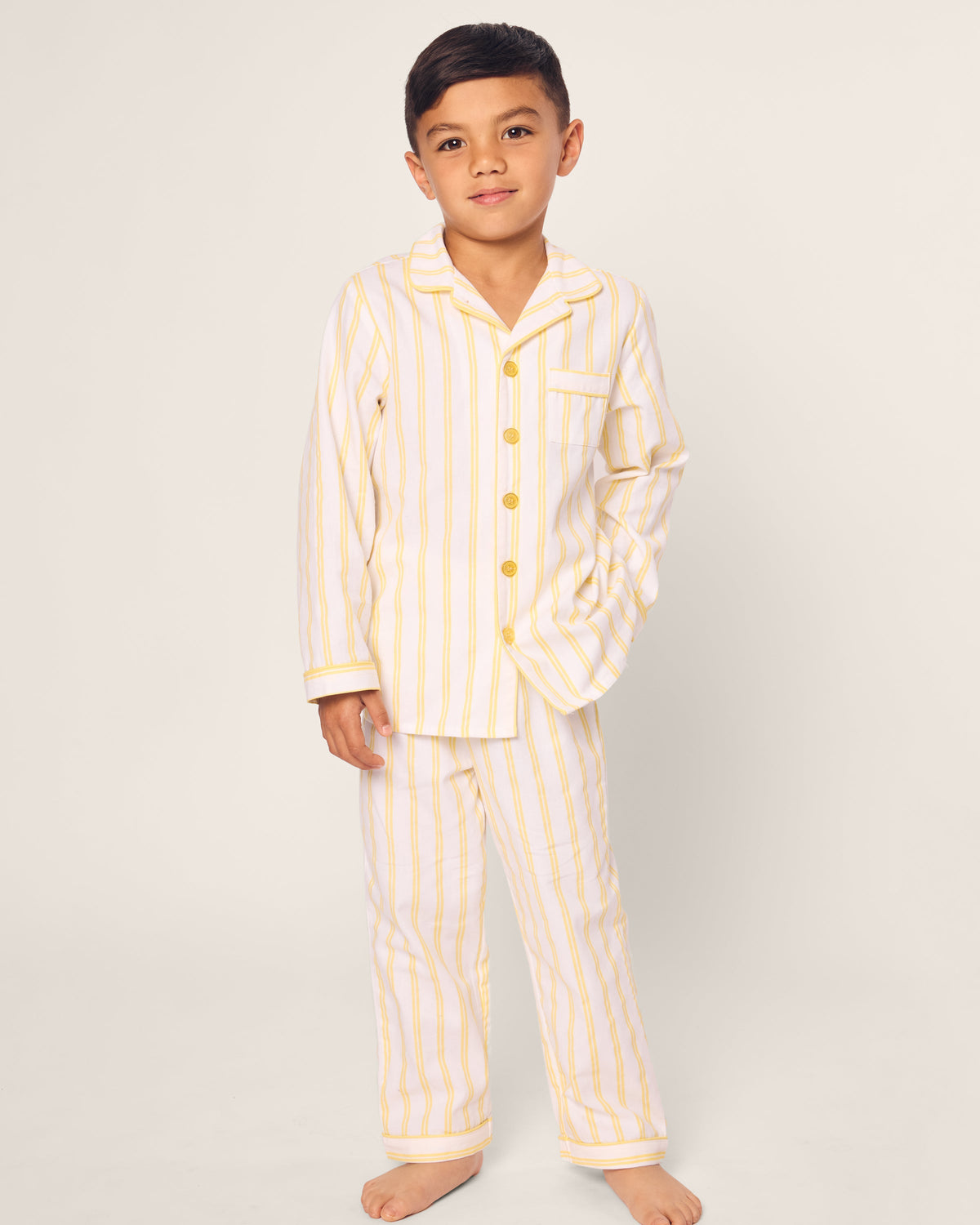 A young boy stands barefoot wearing the Kids Twill Pajama in Sunny Stripe by Petite Plume, showcasing yellow and white stripes with buttons and a shirt pocket. He poses with one hand in his pocket against a plain background.