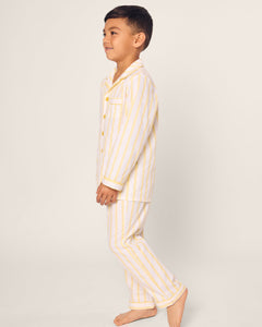 A young boy stands sideways wearing Petite Plumes Kids Twill Pajama in Sunny Stripe, featuring white and yellow vertical stripes. The plain, light-colored background accentuates the cozy design.