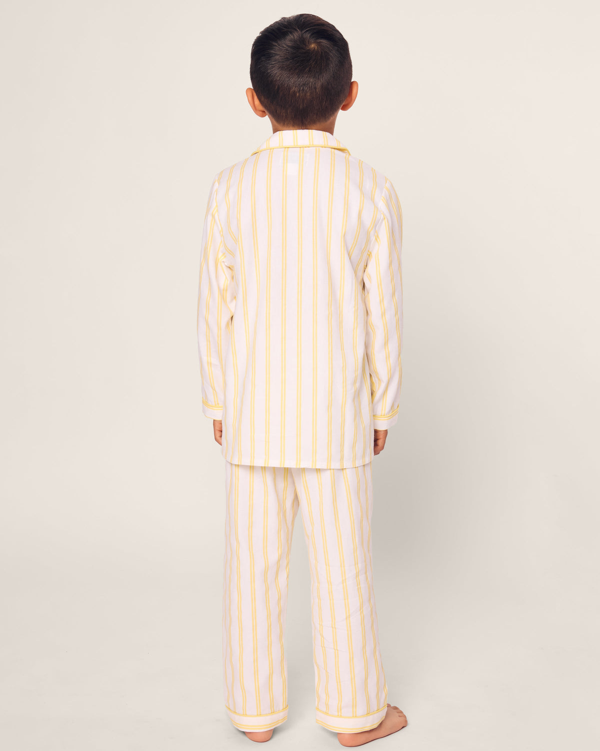 A child wearing Petite Plumes Kids Twill Pajama in Sunny Stripe stands against a light background, highlighting the safety and comfort of the flame-retardant, yarn-dyed cotton with yellow and white vertical stripes.