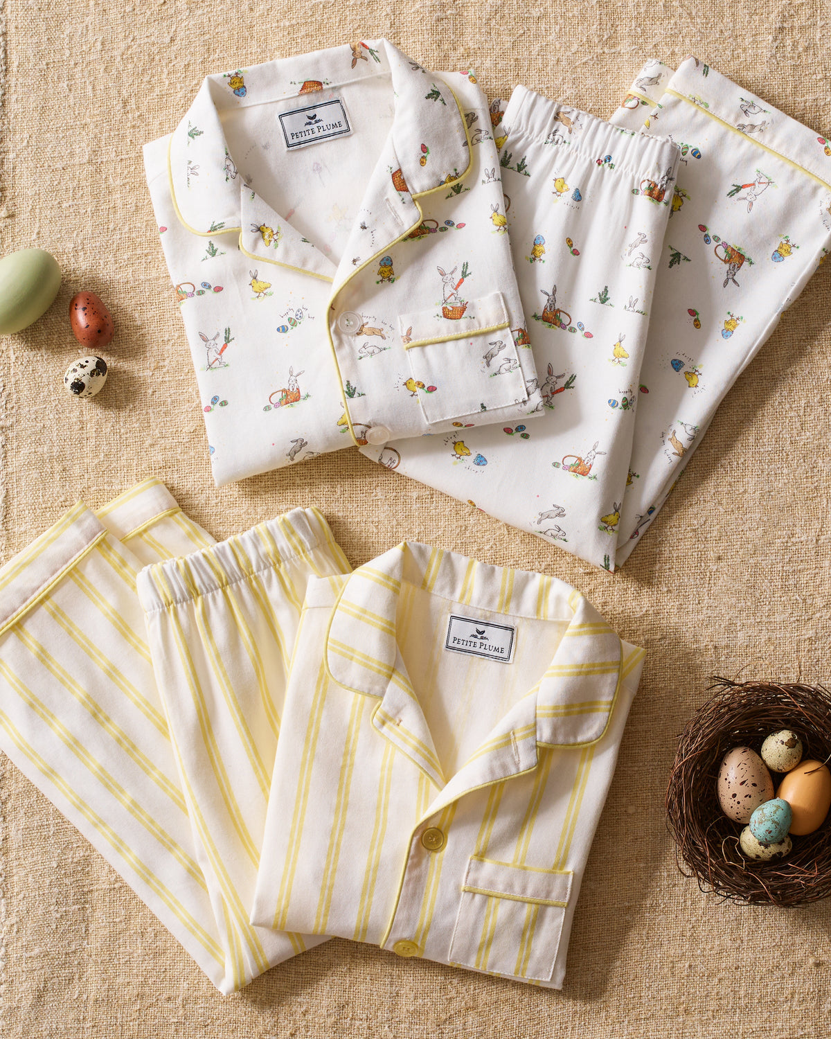 Two sets of Petite Plume childrens pajamas rest on a textured beige surface: one with a fun bunny and flower print, the other is the Kids Twill Pajama in Sunny Stripe. Nearby are small decorative eggs and a nest with speckled eggs.