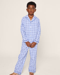 A young boy sports Petite Plumes Kids Twill Pajama Set in Royal Blue Gingham. He is barefoot with short, curly hair, and a plain background highlights his luxury sleepwear’s gingham print.