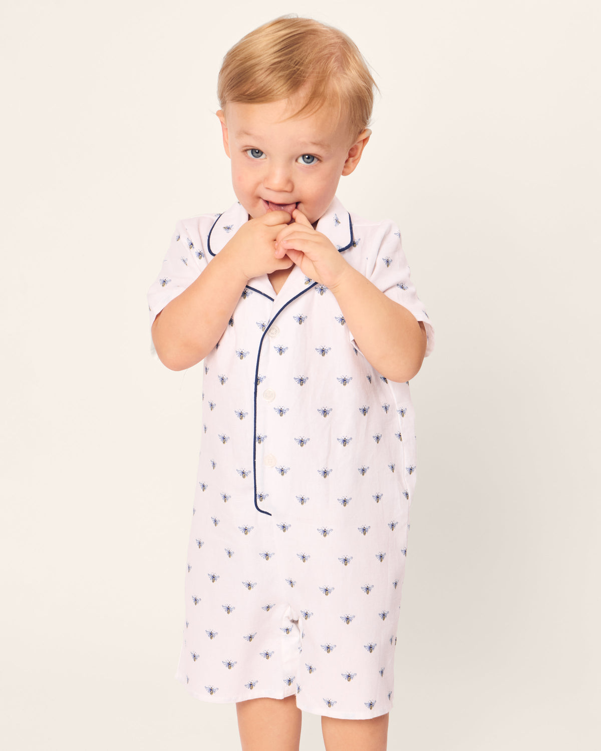 A toddler in Petite Plumes Babys Twill Summer Romper in Buzzing Bees, featuring light-colored brushed twill fabric with bee patterns and dark piping, stands against a plain background, playfully placing fingers in their mouth while gazing at the camera.