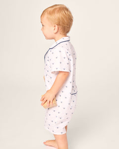 A toddler in a Petite Plume Babys Twill Summer Romper - Buzzing Bees stands on a plain background, holding a block. The lightweight brushed twill fabric ensures comfort, while the childs short blond hair captures the playful summer spirit in profile, showcasing their left side.