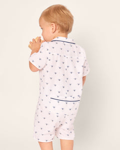 A light-haired toddler faces away, clad in Petite Plumes Babys Twill Summer Romper in Buzzing Bees. The white romper, adorned with navy bee patterns and trim, features the child clapping against a plain light background.