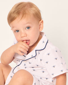 A young child with light hair and blue eyes sits in a Petite Plume Babys Twill Summer Romper in Buzzing Bees, looking thoughtful with fingers near their mouth against a plain background.