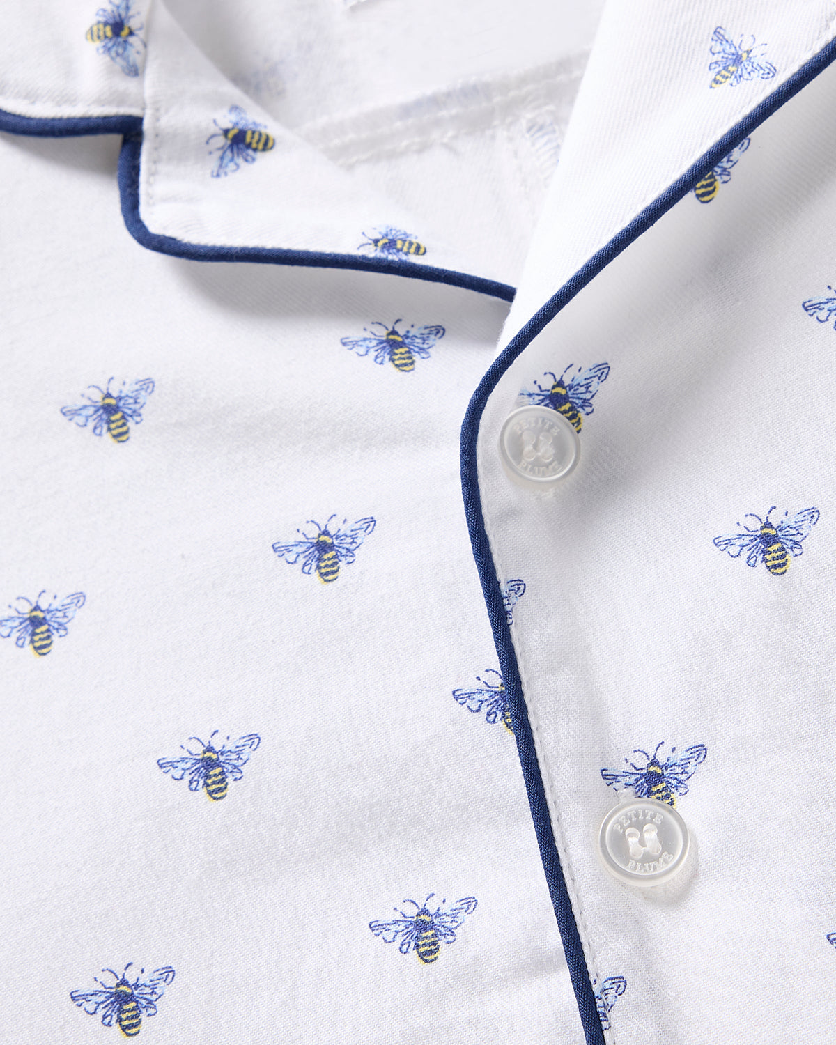 The Babys Twill Summer Romper in Buzzing Bees by Petite Plume features a close-up of white brushed twill with blue and yellow bee patterns, navy blue piping, and white buttons.