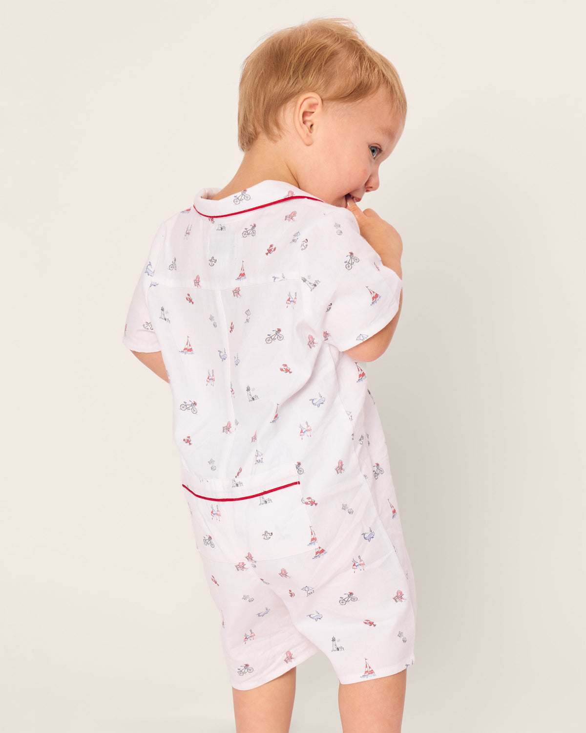 A toddler in Petite Plumes Babys Twill Summer Romper in Coastal Charms, featuring a playful print with bicycles, dogs, and Eiffel Towers, stands slightly turned. The romper is made of brushed twill with red piping accents. The child, gazing aside, has light brown hair.