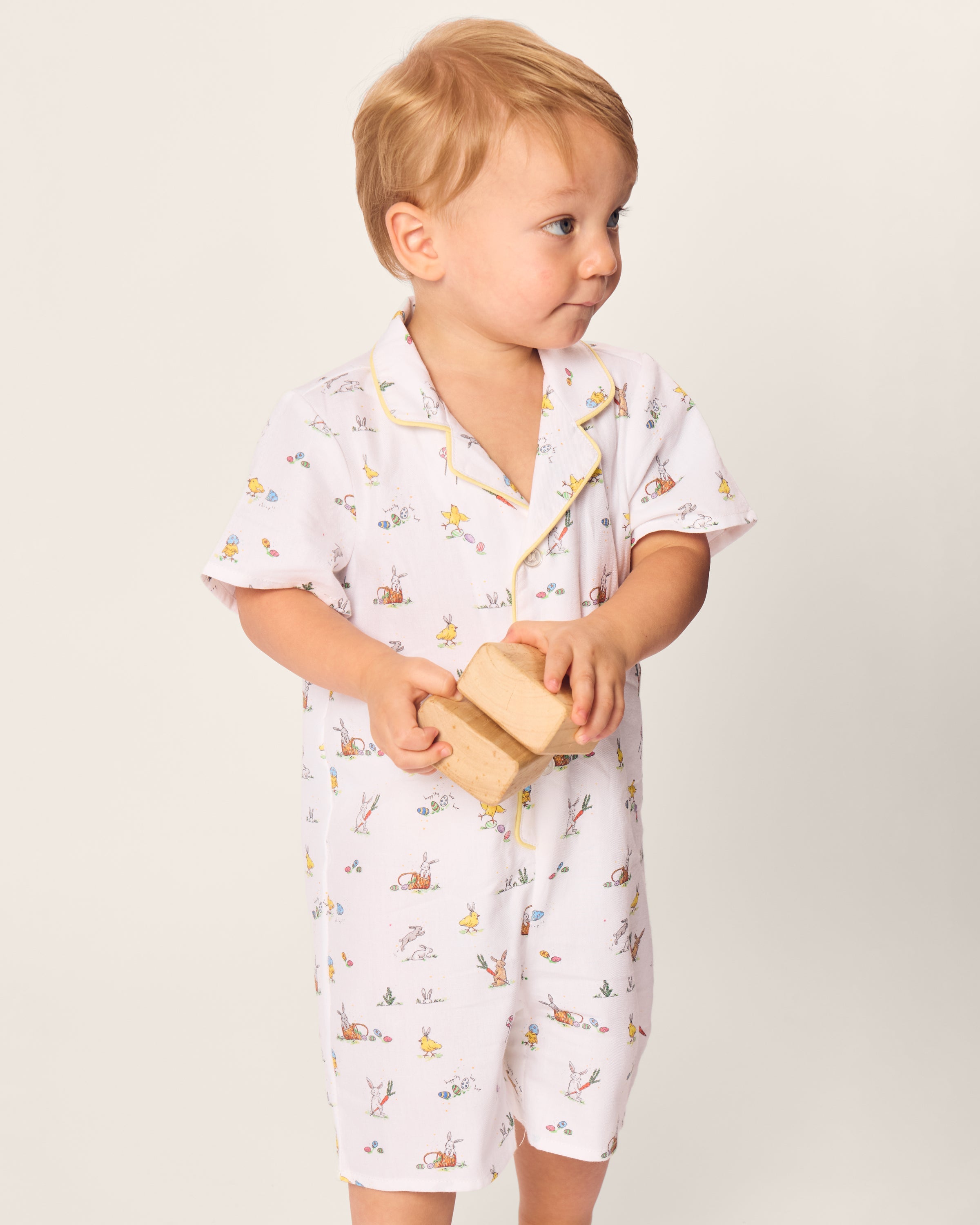 A light-haired toddler wears Petite Plumes Babys Twill Romper in Easter Frolic. The child holds a wooden block, gazing upward. The plain, light background emphasizes the comfort of the brushed twill fabric.