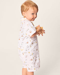 A toddler with light hair, in Petite Plumes Babys Twill Romper in Easter Frolic print, stands sideways, glancing over their shoulder. The romper’s brushed twill fabric complements the plain, light-colored background as the child holds a wooden toy.