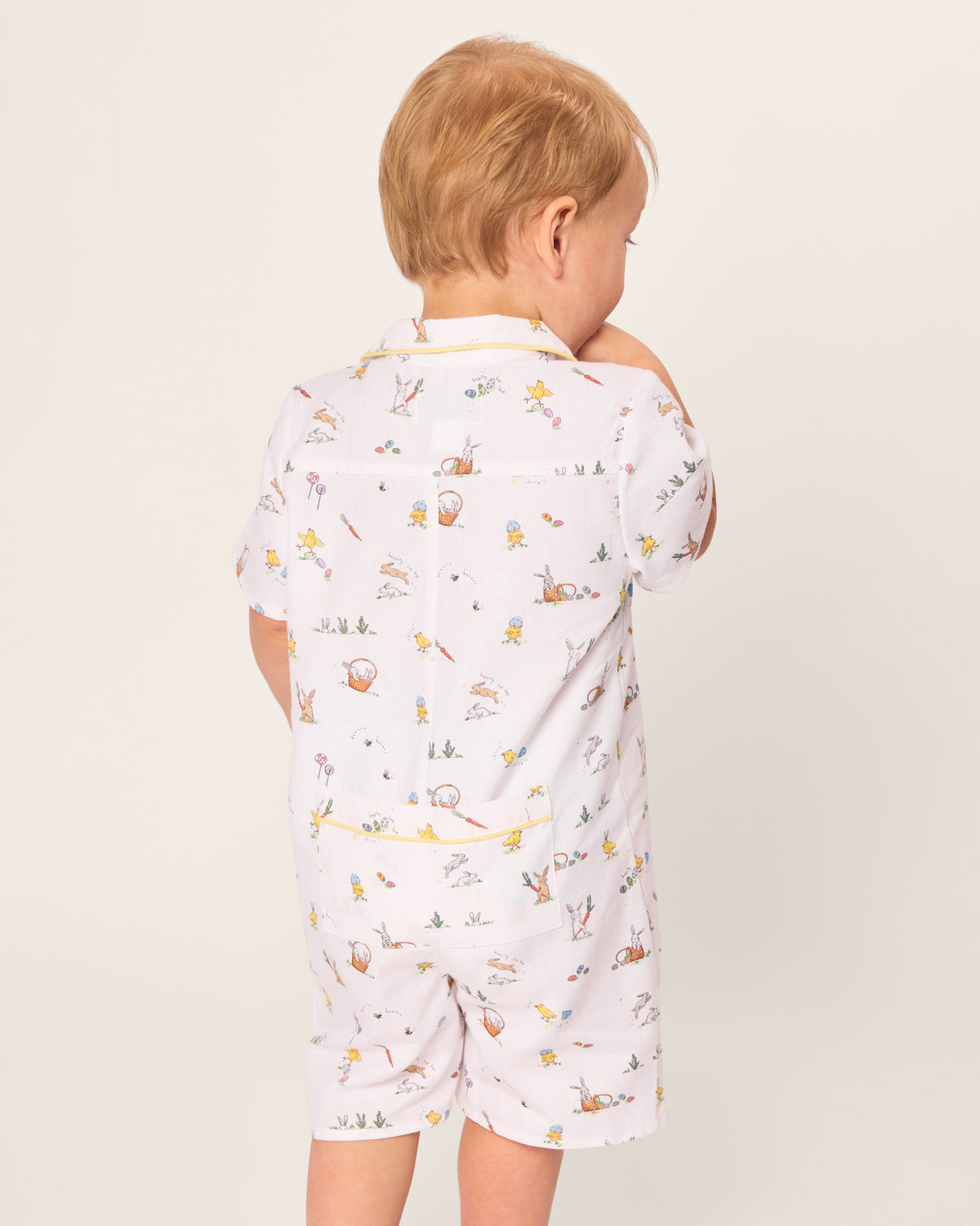 A toddler in Petite Plumes Babys Twill Romper, featuring the Easter Frolic print, gazes to the side. With soft brushed twill fabric showcasing rabbits and gardening tools, the light-haired child enjoys short sleeves and shorts in this charming attire.