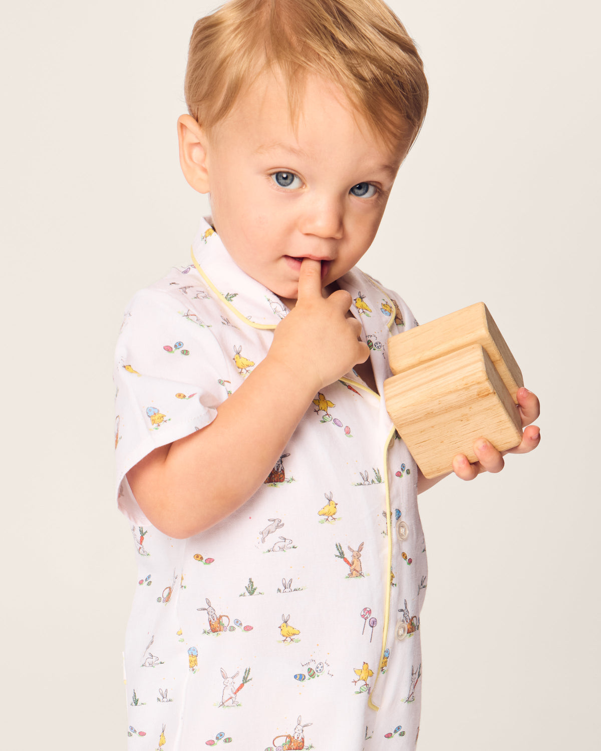 A toddler with light hair, wearing Petite Plumes Babys Twill Romper in Easter Frolic, holds a wooden block and looks at the camera with one finger in their mouth against a plain background.