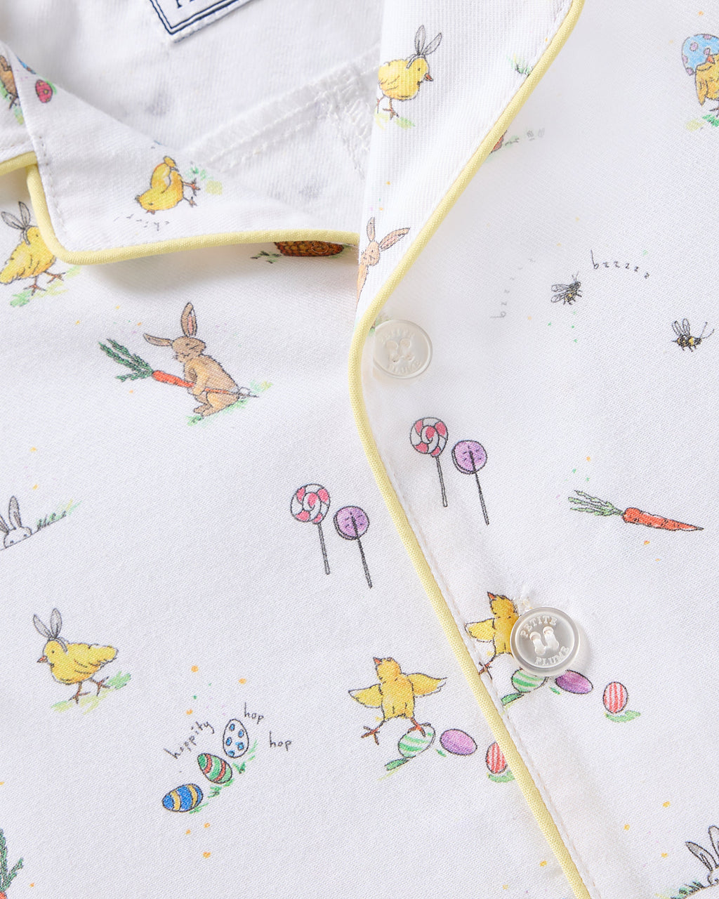 The Babys Twill Romper in Easter Frolic by Petite Plume features a whimsical design of rabbits, chicks, lollipops, and Easter eggs on brushed twill fabric. It includes yellow piping, two white buttons, and is perfect for the season.