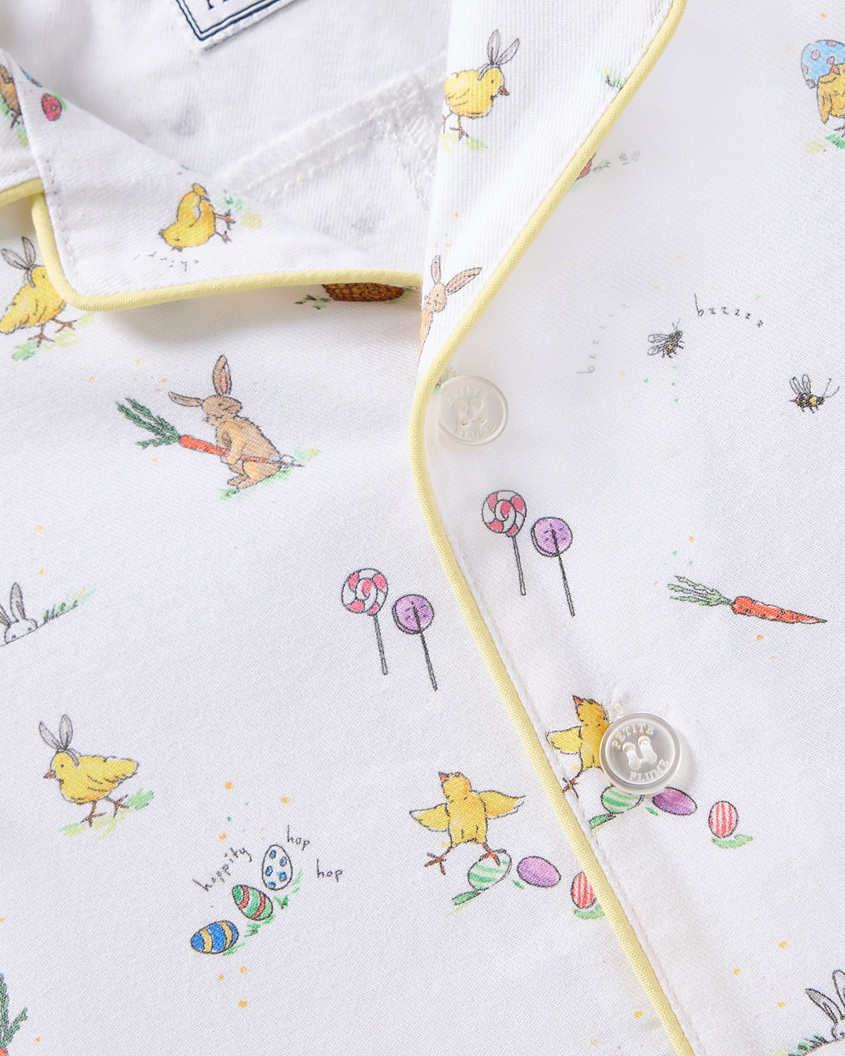 The Babys Twill Romper in Easter Frolic by Petite Plume features a whimsical design of rabbits, chicks, lollipops, and Easter eggs on brushed twill fabric. It includes yellow piping, two white buttons, and is perfect for the season.