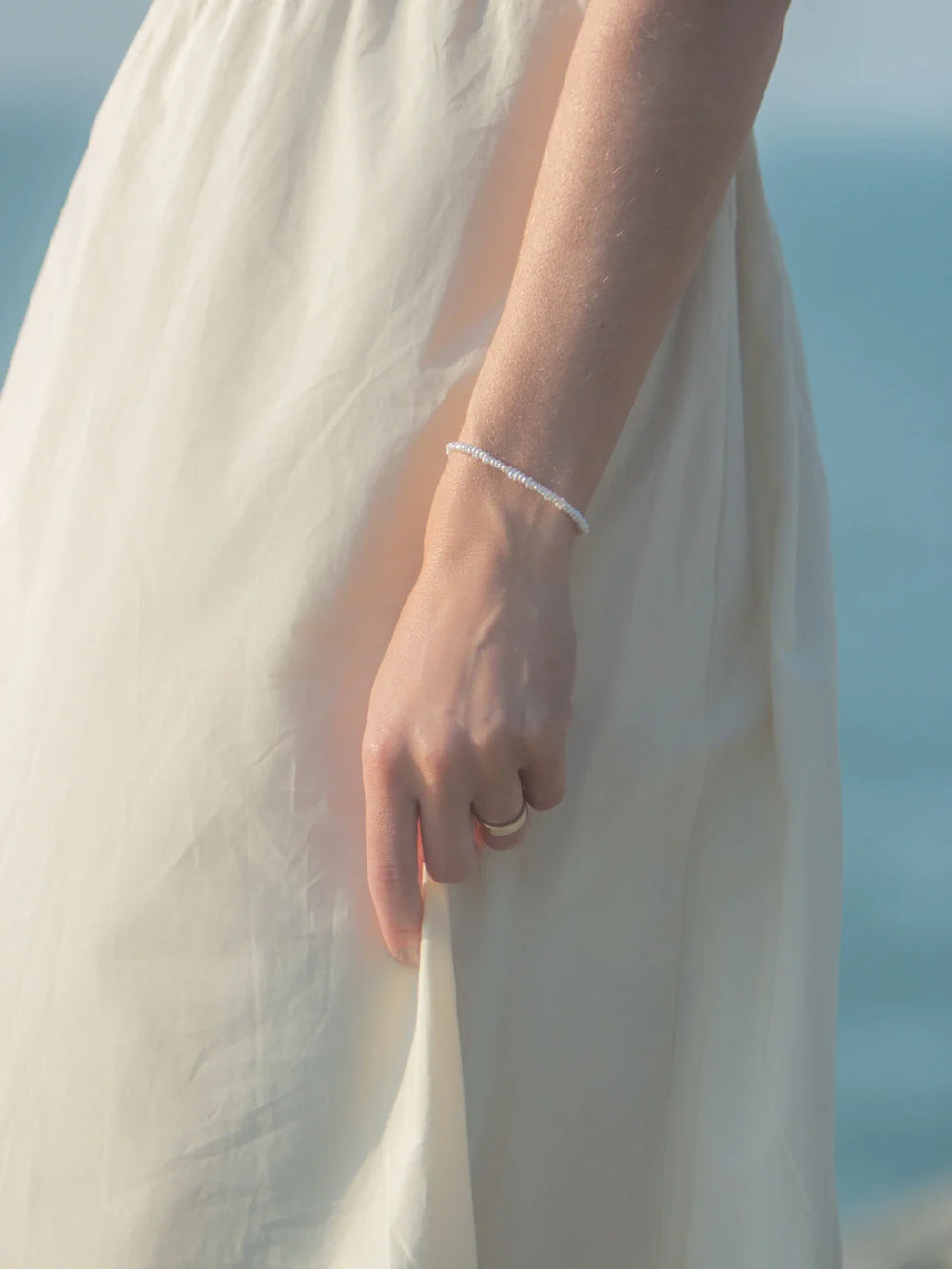 Close-up of Crew Bracelet on a model’s wrist showing small white freshwater pearls against a cream dress and sea background
