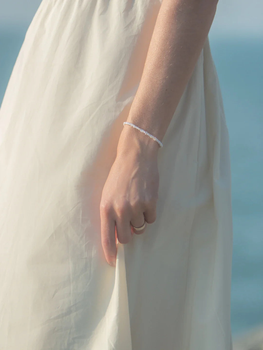 Close-up of Crew Bracelet on a model’s wrist showing small white freshwater pearls against a cream dress and sea background