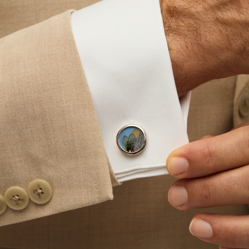 Close-up of Guana cufflink showcasing multicolored natural feathers in rhodium-plated setting on a white shirt cuff and beige jacket