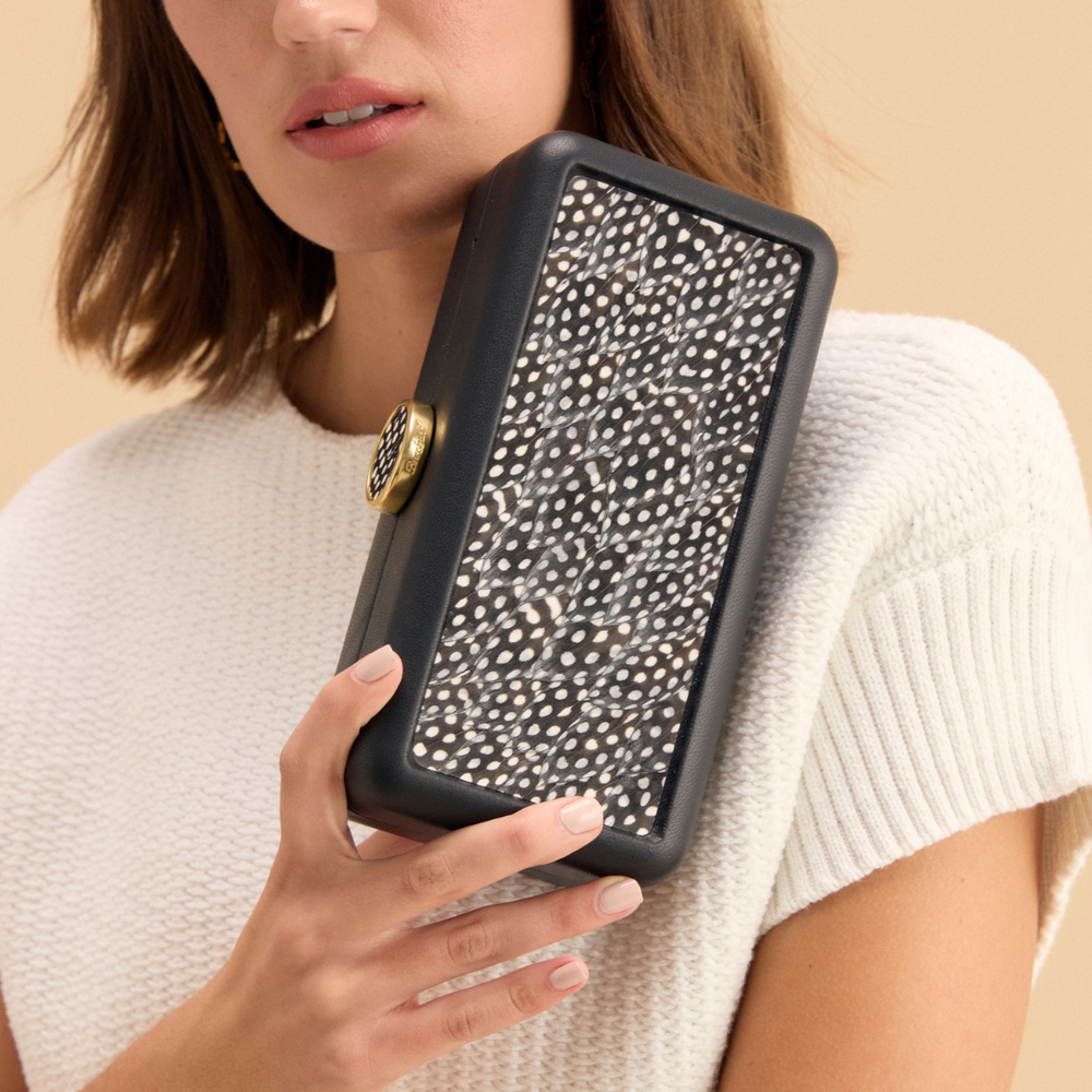 Three-quarter view of Le Marais clutch showing black-and-white guinea feather panel and gold clasp, held by model