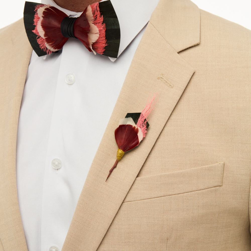 Close-up of Praslin Pin on a beige suit lapel showcasing layered red, coral, and dark green feathers with a gold-wrapped stem