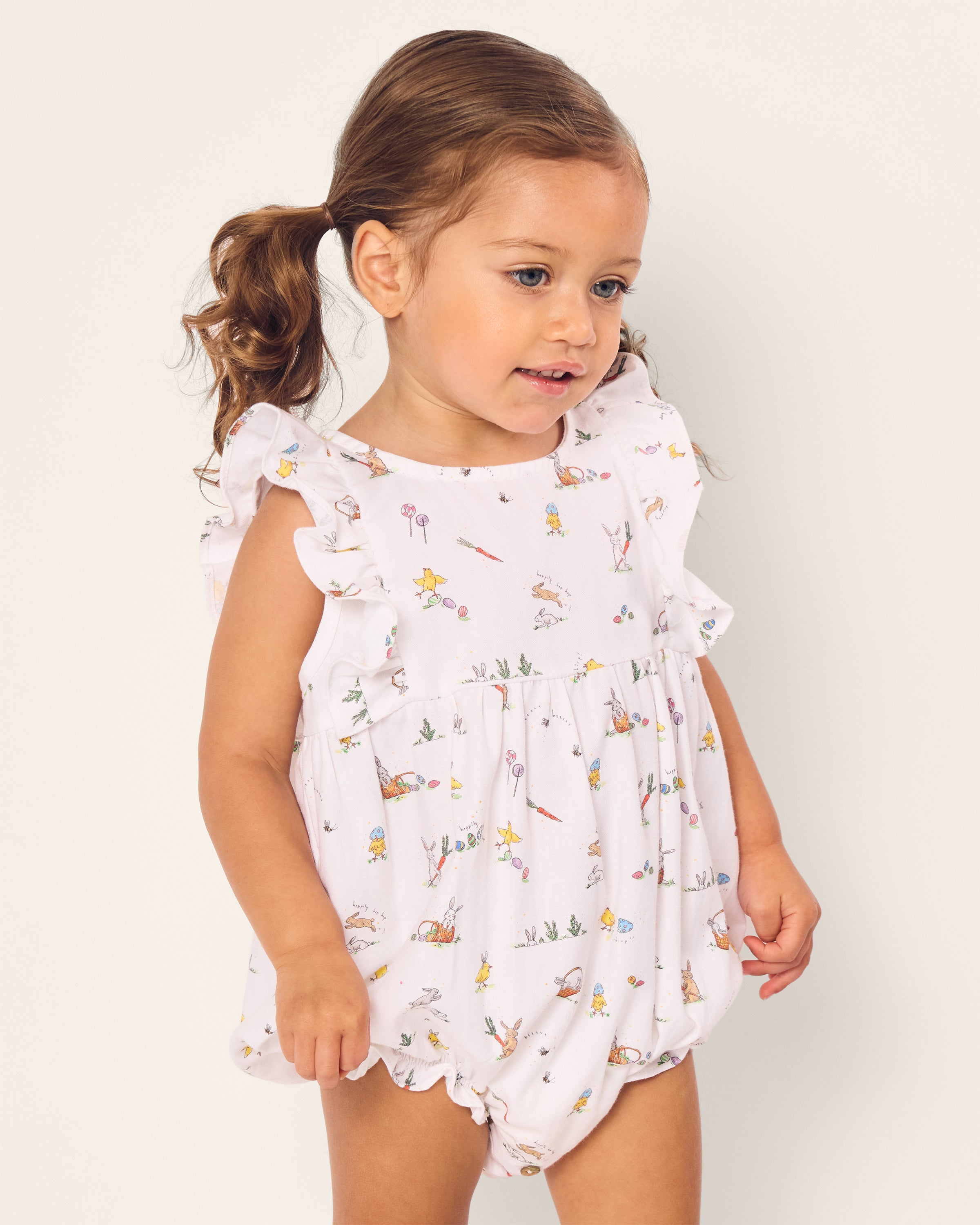 A brown-haired toddler with pigtails wears Petite Plumes Babys Twill Ruffled Romper in Easter Frolic, adorned with ruffled sleeves and vibrant floral and animal patterns, as she gazes left against a plain background.