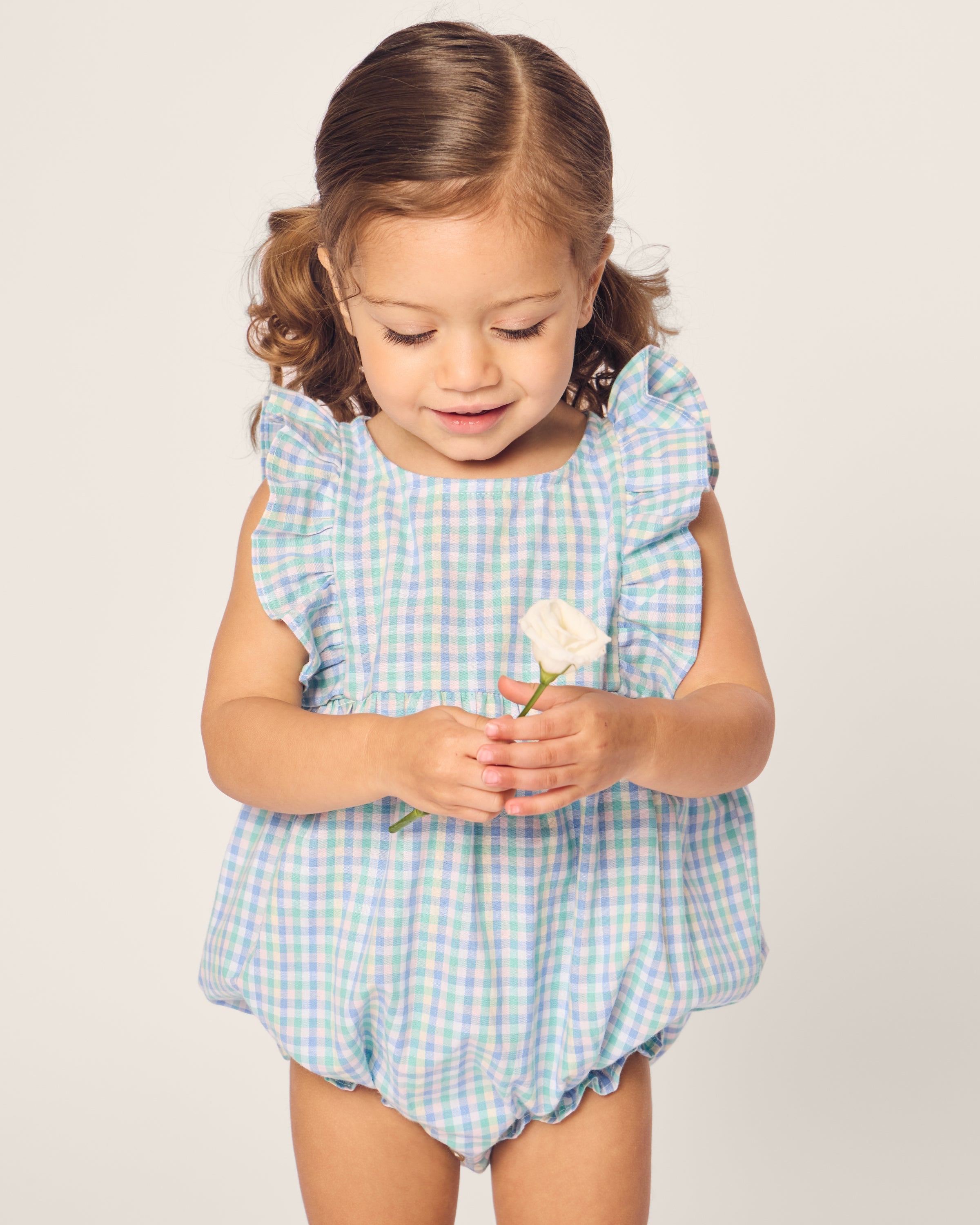 A toddler with pigtails, wearing Petite Plumes Babys Twill Ruffled Romper in Spring Gingham, holds a small white rose and gazes down at it against a plain background.