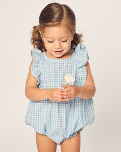 A toddler with pigtails, wearing Petite Plumes Babys Twill Ruffled Romper in Spring Gingham, holds a small white rose and gazes down at it against a plain background.