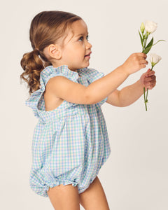 A young child in Petite Plumes Babys Twill Ruffled Romper in Spring Gingham, holding white flowers and wearing a blue-green checkered outfit with ruffled sleeves, gazes upward curiously against a soft beige background.