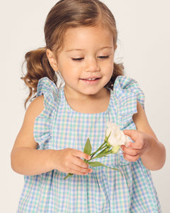 A young girl with brown pigtails smiles gently while holding a white flower. She is wearing Petite Plumes Babys Twill Ruffled Romper in Spring Gingham against a plain background.