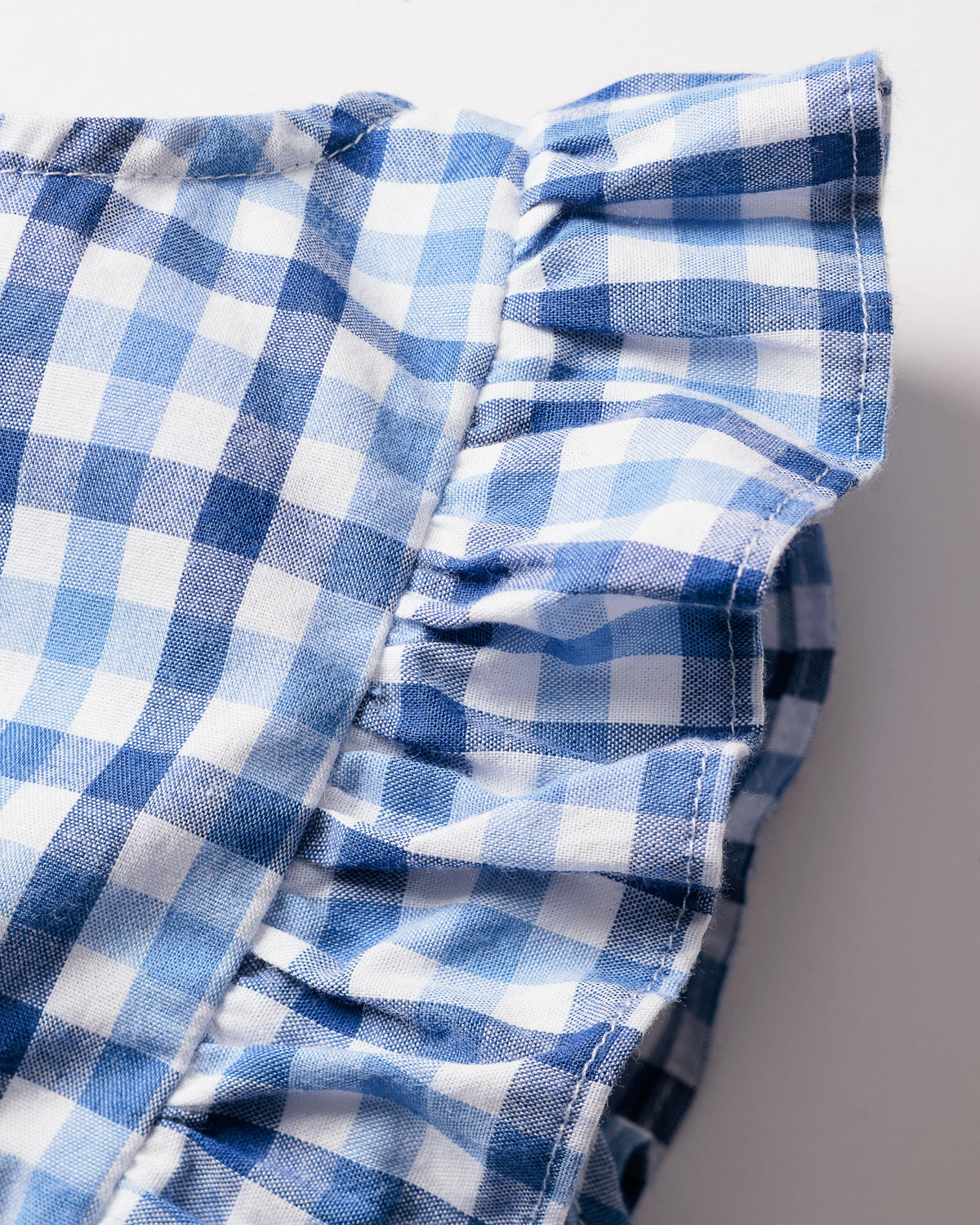 A close-up shows the Babys Twill Ruffled Romper in Royal Blue Gingham by Petite Plume, featuring a classic gingham print with alternating light and dark blue squares. The fabrics slightly wrinkled texture complements its brushed material and ruffled edges.