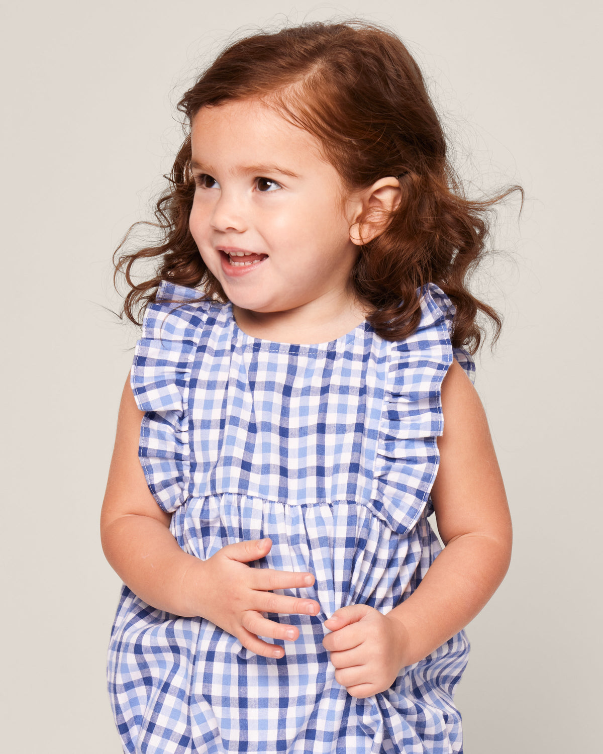 A young child with wavy brown hair wears Petite Plumes Babys Twill Ruffled Romper in Royal Blue Gingham, smiling and looking sideways against a plain background.