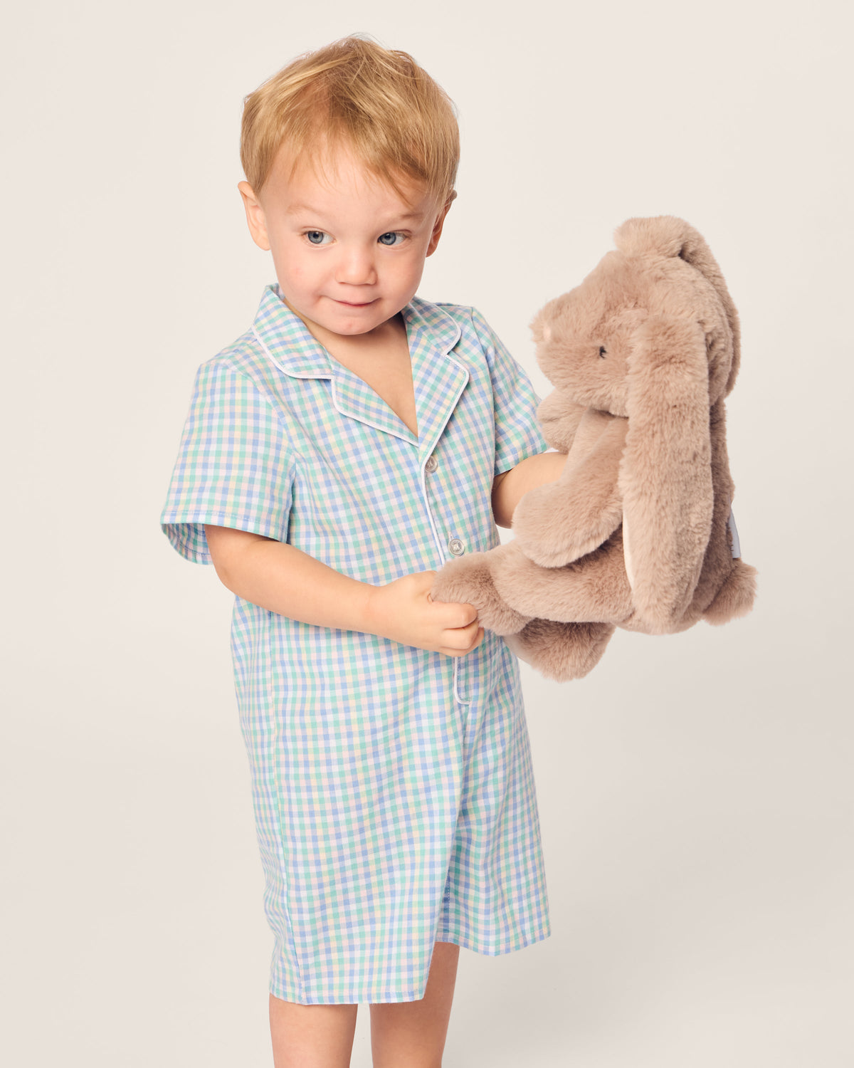 A young child with light brown hair wears a pastel-colored Babys Twill Romper in Spring Gingham by Petite Plume. They hold a plush brown bunny with long ears, looking happy against the plain, light-colored background.