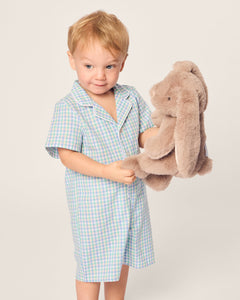 A young child with light brown hair wears a pastel-colored Babys Twill Romper in Spring Gingham by Petite Plume. They hold a plush brown bunny with long ears, looking happy against the plain, light-colored background.