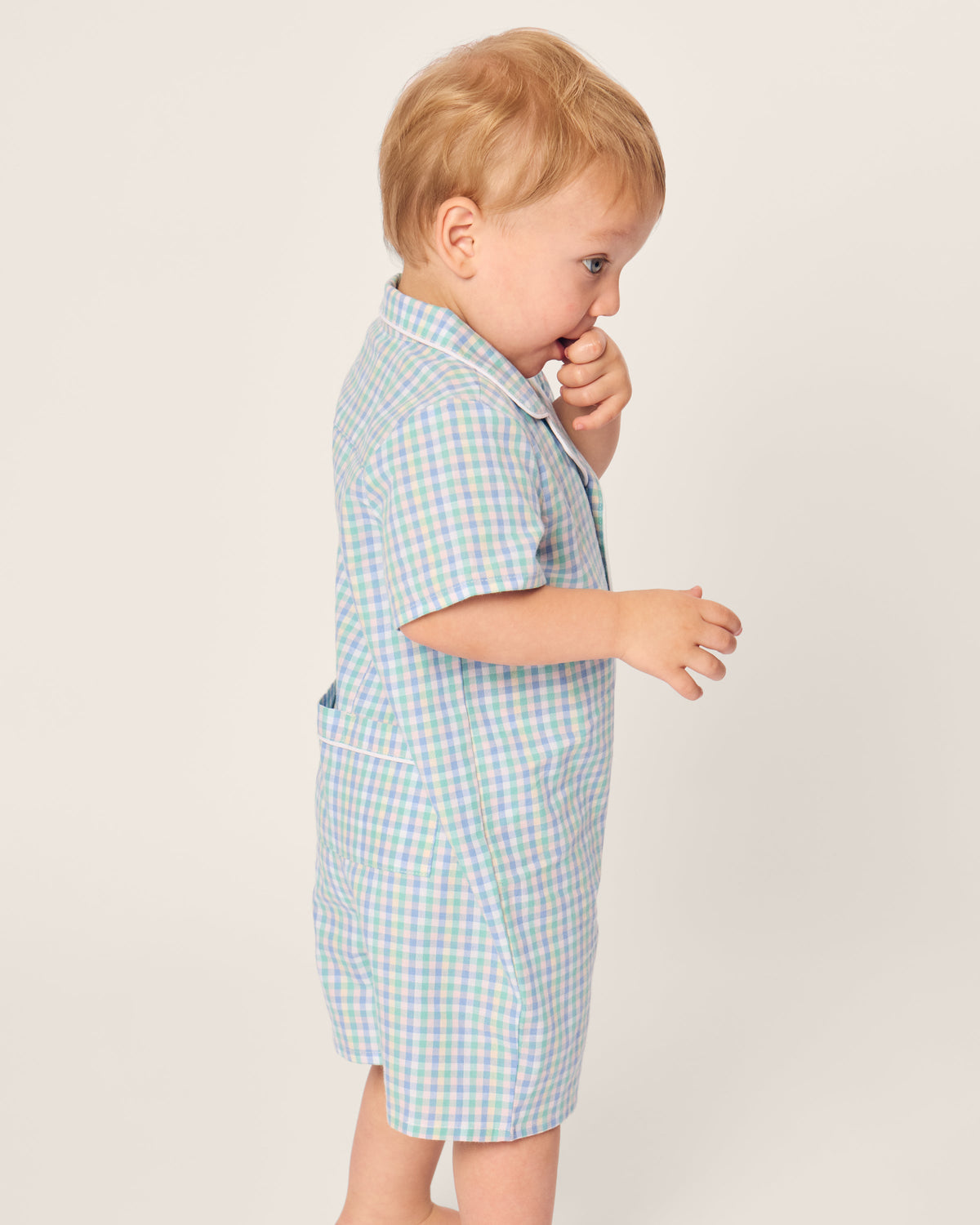 A toddler with light hair, dressed in a charming Petite Plume Babys Twill Romper in Spring Gingham, stands sideways facing left, raises one hand to their mouth, and gazes slightly off to the side against a plain off-white background.