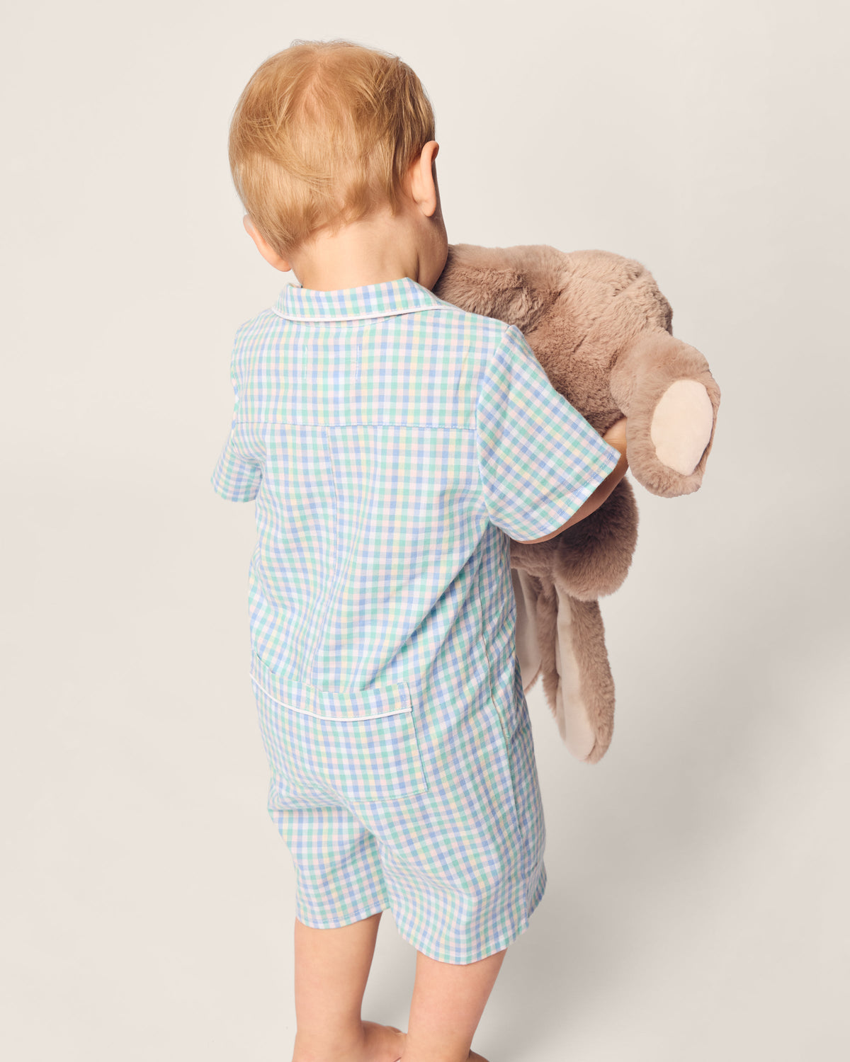 A toddler with light brown hair, seen from behind on a light gray background, wears the Petite Plume Babys Twill Romper in Spring Gingham, a charming blue and green checkered romper made from soft yarn-dyed cotton, while holding a large brown stuffed bear.