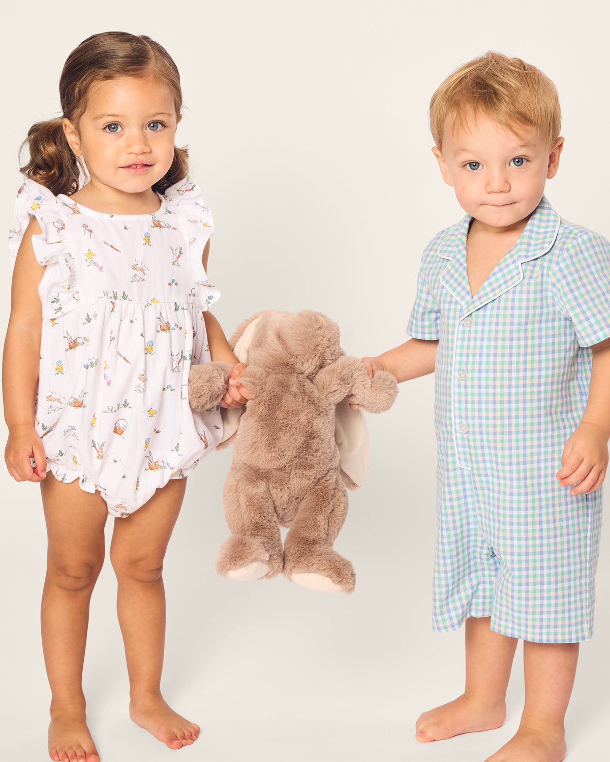 A girl in a floral summer romper and a boy wearing Petite Plumes Babys Twill Romper in Spring Gingham stand holding a brown plush bunny, both looking at the camera with neutral expressions against a plain off-white background.