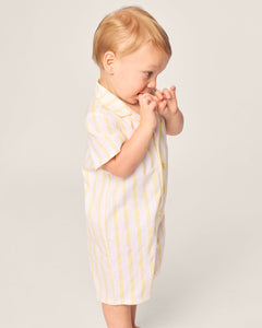 A toddler with blond hair stands sideways in a Petite Plumes Babys Twill Summer Romper in Sunny Stripe, fingers near the mouth against a plain light-colored background.