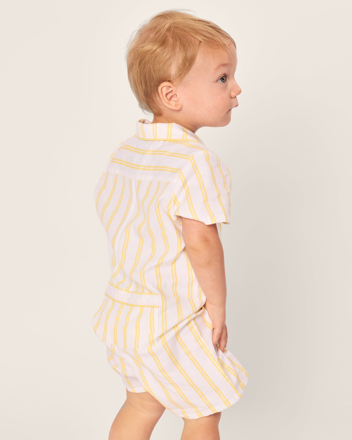 A toddler with blonde hair is wearing a Petite Plume Babys Twill Summer Romper in Sunny Stripe. The romper features yarn-dyed cotton with yellow and white stripes, as the child looks to the side against a plain, light background.