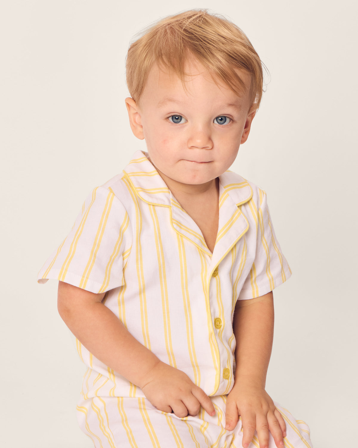 A young child with short light hair sits wearing Petite Plumes Babys Twill Summer Romper in Sunny Stripe, made from yarn-dyed cotton with white and yellow vertical stripes. The plain off-white background enhances the soft, summery atmosphere.