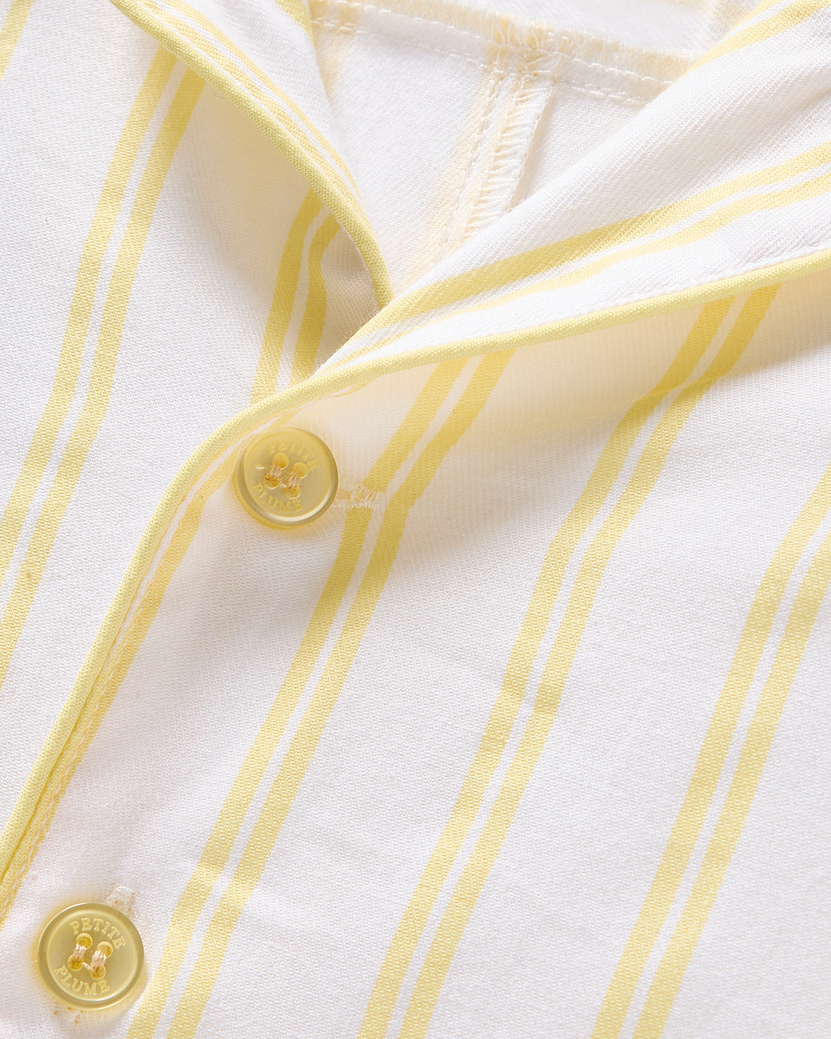 Close-up of Petite Plumes Babys Twill Summer Romper in Sunny Stripe, showcasing white brushed twill with light yellow vertical stripes and two four-hole pale yellow buttons, slightly unbuttoned to reveal neat stitching that enhances its elegant soft texture.